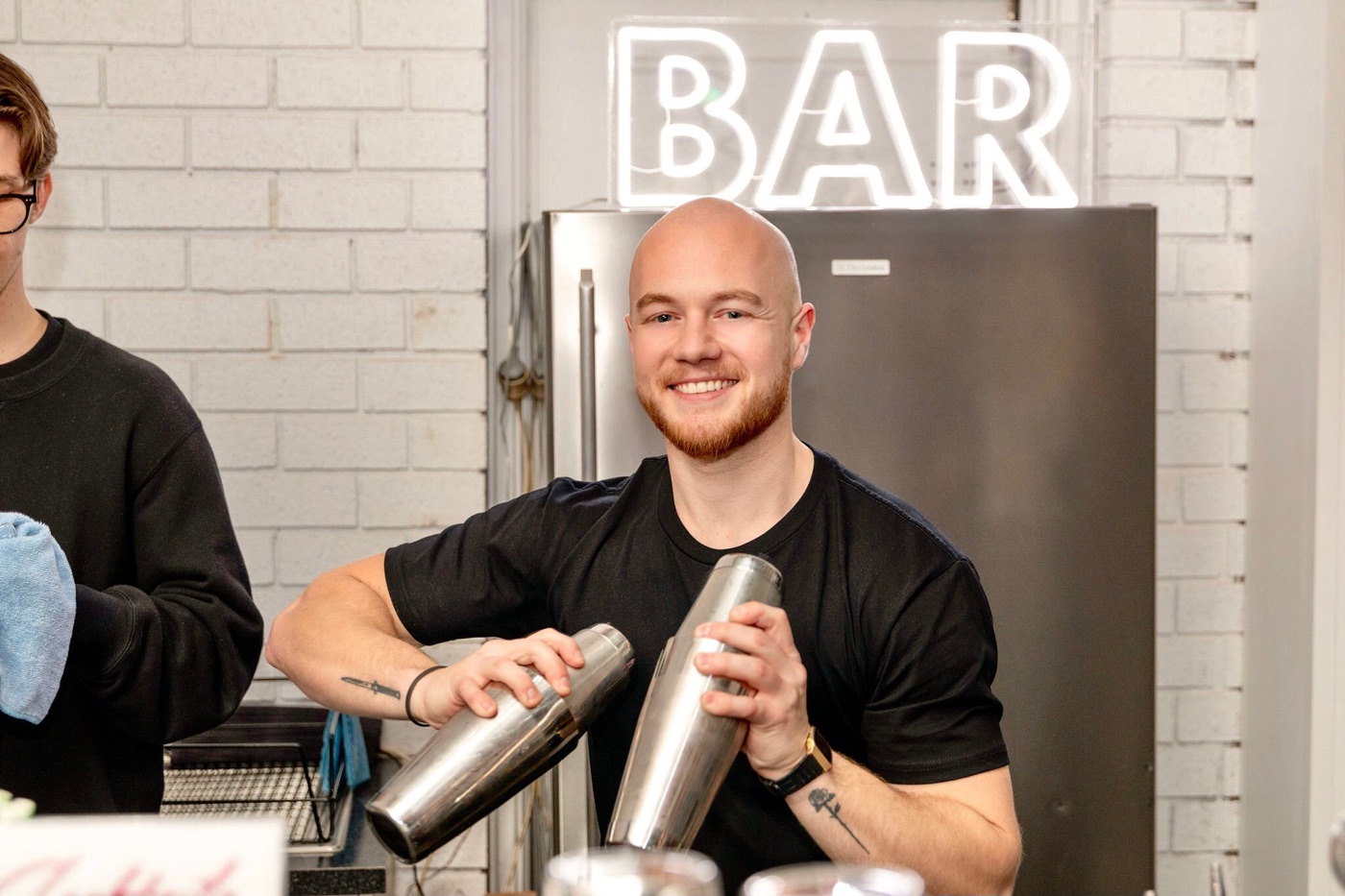 Scott, the Scottails cocktail teacher, shaking two tins under a neon bar sign